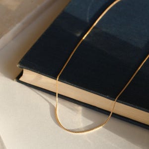 May include: A delicate gold-colored chain necklace resting on a dark blue book. The chain has a fine, box-style link design. The book is closed, with a textured cover. The scene is lit with soft, natural light, creating subtle shadows.