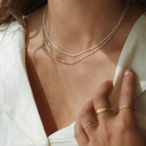 May include: Two delicate necklaces, one gold and one silver, rest on a person's neck. The gold necklace is at the bottom, and the silver necklace is above it. Both necklaces have small, evenly spaced beads. Several gold rings adorn the fingers.