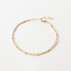 以下が含まれることがあります： 繊細なゴールドトーンのブレスレット。2本の平行なチェーンがあり、1本は小さなビーズが等間隔に並び、もう1本は細かいテクスチャのデザイン。ブレスレットは調節可能な留め金付きで、白い背景に配置されています。