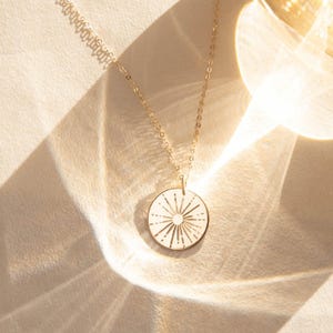 May include: A gold-colored necklace with a circular pendant. The pendant features a sunburst design with etched lines radiating from the center. The necklace chain is delicate and the pendant hangs on a white surface.