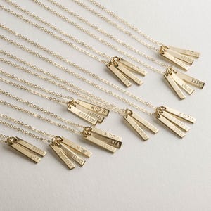 Könnte beinhalten: Goldfarbene Halsketten mit rechteckigen Anhängern. Jeder Anhänger ist mit Namen und Symbolen graviert. Die Halsketten werden auf hellem Hintergrund präsentiert und zeigen den personalisierten Schmuck.