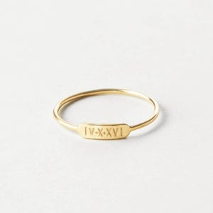 Op de afbeelding: Een delicate gouden ring met een rechthoekige plaat met de Romeinse cijfers IV-X-XVI. De ring is dun en minimalistisch. De ring wordt gepresenteerd tegen een witte achtergrond.