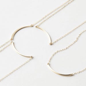 May include: Gold-toned necklaces with a delicate chain and a curved bar pendant. The necklaces are displayed on a white surface, showcasing their minimalist design. The curved bar pendant is approximately 1 inch in length.