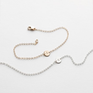 May include: Two delicate chain bracelets. One is gold-toned with a circular charm engraved with the letter "A". The other is silver-toned with a circular charm engraved with the letter "K". Both bracelets have a lobster clasp closure.