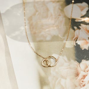 May include: A delicate gold necklace featuring two interlocking circles. The necklace has a fine chain and a simple, elegant design. The circles are of different sizes, with one slightly smaller than the other. The jewelry is displayed on a floral background.