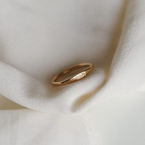 May include: A gold-colored ring with a delicate leaf design. The ring is set against a soft, off-white fabric, highlighting its polished surface and intricate details. The ring's band is smooth and rounded.