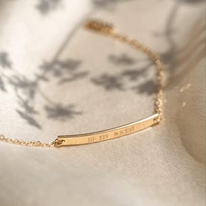 Puede incluir: Una delicada pulsera dorada con un colgante de barra. La barra está inscrita con números romanos. La pulsera está sobre una superficie suave y clara, con sombras en el fondo. Es una joya.