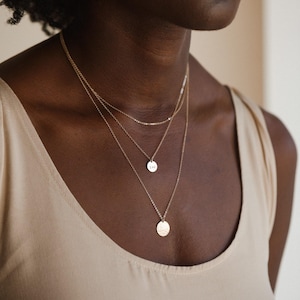 May include: A layered gold necklace set. The necklaces are of varying lengths, with a delicate chain and a small circular pendant. The bottom necklace has a textured, hammered design. The model is wearing a beige tank top.