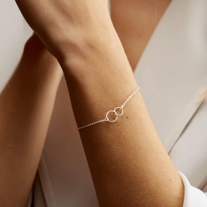 May include: A silver bracelet with two interlinked circles. The bracelet is attached to a fine chain, creating a minimalist design. This piece of jewellery is suitable for everyday wear or special occasions.
