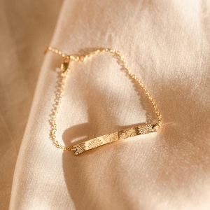 May include: A gold-colored bar bracelet with a delicate chain. The bar features an engraved floral design. The bracelet is displayed on a satin, beige surface, creating a soft, elegant aesthetic. The bracelet is a piece of jewelry.