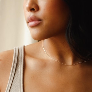 May include: A delicate gold necklace resting on a person's collarbone. The fine chain is a simple, elegant design. The person is wearing a neutral-colored tank top.