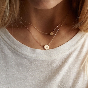 Puede incluir: Dos delicados collares dorados, superpuestos, cada uno con un pequeño colgante redondo. El colgante inferior muestra la letra "K", mientras que el colgante superior muestra la letra "A". Los collares se llevan sobre una camiseta blanca.