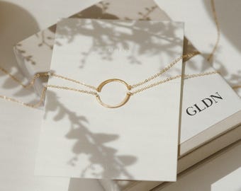 May include: A delicate gold necklace with a circular pendant is displayed on a white card. The necklace is placed on a box with the letters "GLDN" printed on it. The image is lit with soft, natural light.