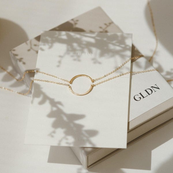 Pode incluir: Um delicado colar de ouro com um pingente circular é exibido em um cartão branco. O colar é colocado em uma caixa com as letras "GLDN" impressas. A imagem é iluminada com luz suave e natural.