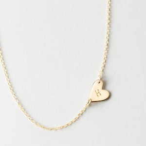 Könnte beinhalten: Eine zarte, goldfarbene Halskette mit einem herzförmigen Anhänger. Der Anhänger trägt den eingravierten Buchstaben "H". Die Kette hat ein feines, gegliedertes Design, und der Hintergrund ist eine weiche, neutrale Farbe.