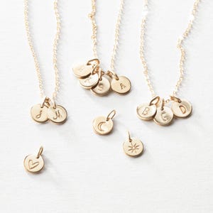 May include: Gold-colored necklaces with delicate chains and small, round pendants. Some pendants feature engraved initials, while others display a heart, a crescent moon, and a starburst design. The necklaces are displayed on a white surface.