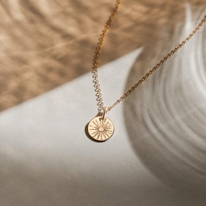 May include: A gold necklace with a circular pendant. The pendant has a sunburst design with etched lines. The delicate chain is displayed on a neutral background. The necklace is a simple, elegant piece.
