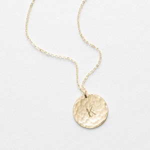 Könnte beinhalten: Goldfarbene Halskette mit einem Anhänger mit Hammerschlag-Textur. Der runde Anhänger trägt den Buchstaben "K" eingraviert. Die Kette ist filigran und goldfarben, vor weißem Hintergrund.