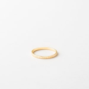 May include: A delicate gold ring with a thin band. The ring features a subtle, repeating pattern of small shapes around the band. The ring is set against a plain white background, highlighting its simple elegance.