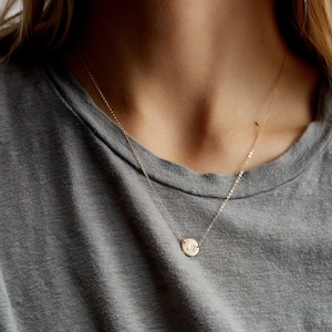 Può includere: Una delicata collana color oro con un piccolo pendente rotondo. Il pendente presenta un'iniziale incisa, sospesa da una sottile catena. La collana è indossata su una maglietta grigia.