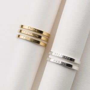 May include: Close-up of two sets of personalized rings. One set is gold-toned with the names "EMMA", "GRAYSON", and "CHLOE" engraved. The other set is silver-toned with "FELICITY" and "HARRIS" engraved. Rings are displayed on white fabric.