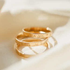 May include: Three gold rings stacked on top of each other. The top ring has the letter "A" and a leaf design. The middle ring has star and crescent moon designs. The rings are on a white, textured surface.