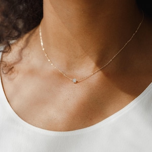 Puede incluir: Un delicado collar de oro con un pequeño colgante de piedra preciosa de color claro. El collar se lleva alrededor del cuello, mostrando el diseño minimalista. La cadena es fina y el colgante es una pequeña perla.