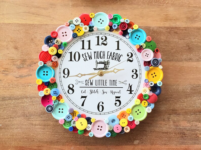 May include: A white clock with black numbers and hands, surrounded by colorful buttons. The clock face reads "Sew Much Fabric" and "Sew Little Time".