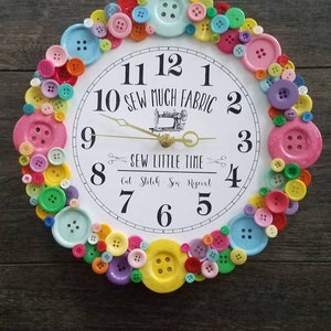 May include: A white clock with black numbers and a sewing machine design. The clock is surrounded by colorful buttons. The text on the clock reads "10 Sew Much Fabric" and "Sew Little Time Cut Stitch Sew Repeat".