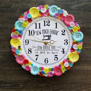May include: A white clock with black numbers and a sewing machine design in the center. The clock is surrounded by a ring of colorful buttons.