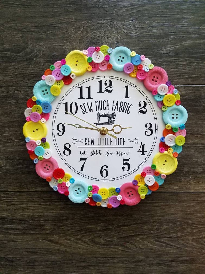 May include: A white clock with black numbers and a sewing machine design in the center. The clock is surrounded by a ring of colorful buttons.