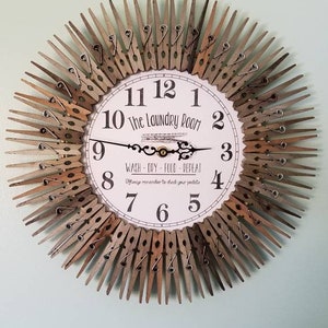 Puede incluir: Un reloj blanco con n&uacute;meros y manecillas negros, rodeado de pinzas de ropa de madera. La esfera del reloj dice "The Laundry Room" y "Wash - Dry - Fold - Repeat".