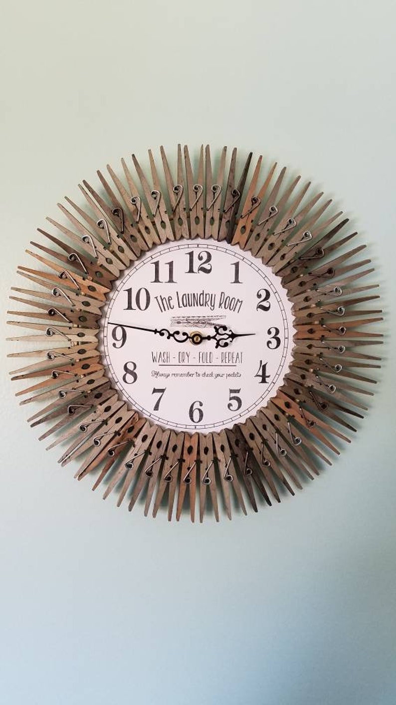 Puede incluir: Un reloj blanco con n&uacute;meros y manecillas negros, rodeado de pinzas de ropa de madera. La esfera del reloj dice "The Laundry Room" y "Wash - Dry - Fold - Repeat".