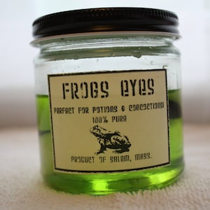 May include: A glass jar with a black lid filled with a green liquid. The label reads "FROGS EYES" and "PERFECT FOR POTIONS & CONCOCTIONS". It also says "100% PURE" and "PRODUCT OF SALEM, MASS.". A black silhouette of a frog is on the label.