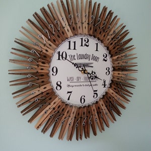 Puede incluir: Un reloj hecho de pinzas de madera con una cara blanca que dice "The Laundry Room" y "Wash - Dry - Fold Repeat" con un recordatorio para revisar los bolsillos. Las manecillas del reloj son negras y los n&uacute;meros son negros.