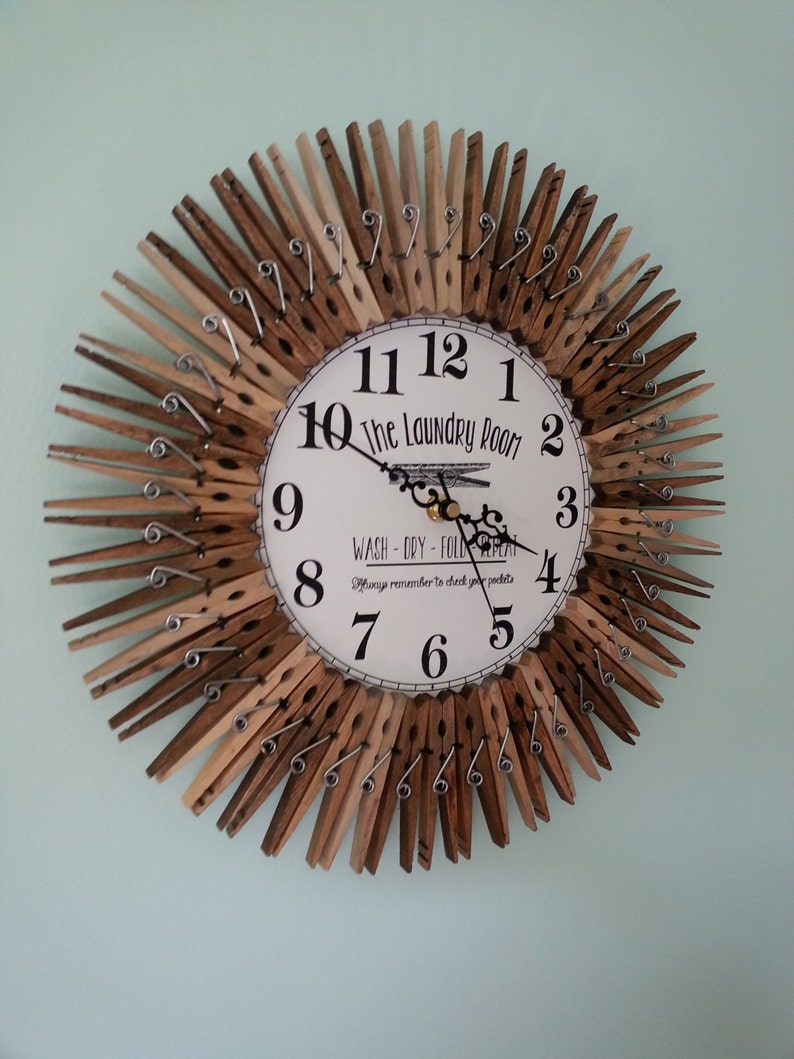 Puede incluir: Un reloj hecho de pinzas de madera con una cara blanca que dice "The Laundry Room" y "Wash - Dry - Fold Repeat" con un recordatorio para revisar los bolsillos. Las manecillas del reloj son negras y los n&uacute;meros son negros.