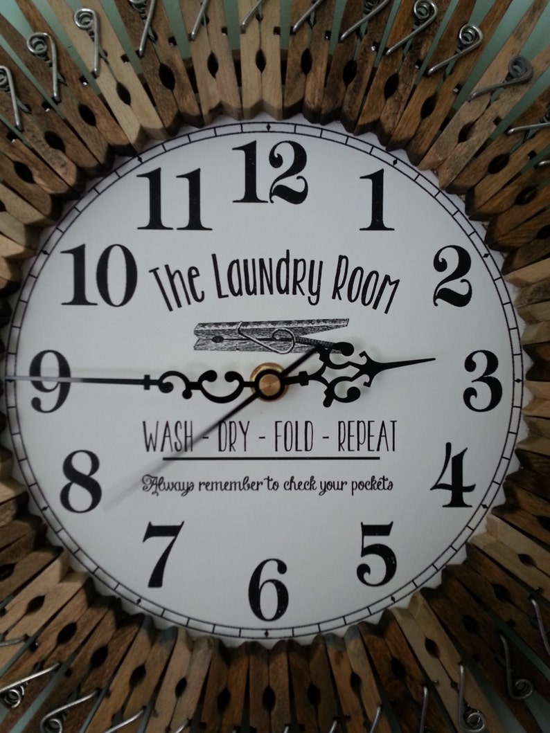 Puede incluir: Un reloj blanco con n&uacute;meros y manecillas negros, rodeado de pinzas de ropa de madera. La esfera del reloj dice "The Laundry Room" y "Wash - Dry - Fold - Repeat". El texto "Always remember to check your pockets" est&aacute; impreso debajo de la esfera del reloj.
