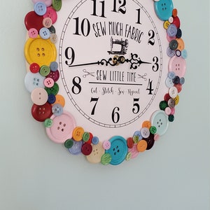May include: A white clock with black numbers and a sewing machine design. The clock is surrounded by colorful buttons.