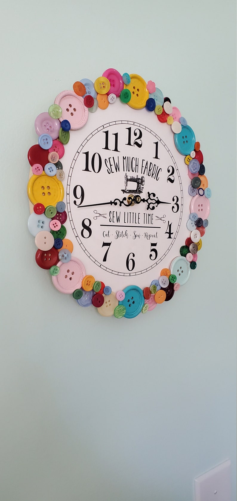May include: A white clock with black numbers and a sewing machine design. The clock is surrounded by colorful buttons.