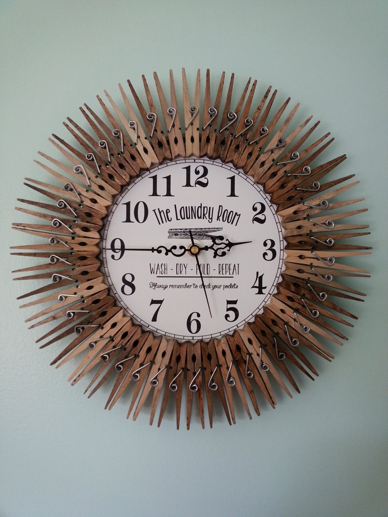 Puede incluir: Un reloj hecho de pinzas de madera con una cara blanca que dice "The Laundry Room" y "Wash - Dry - Fold - Repeat" con un recordatorio para revisar tus bolsillos. Las manecillas del reloj son negras y doradas.