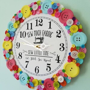 May include: A white clock with a black face and black numbers. The clock is decorated with colorful buttons around the edge. The clock face says "Sew Much Fabric" and "Sew Little Time".