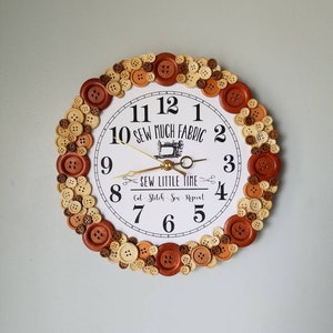 May include: A white clock with brown and beige buttons surrounding the face. The clock face has black numbers and the text "Sew Much Fabric" and "Sew Little Time Cut - Stitch - Sew - Repeat".