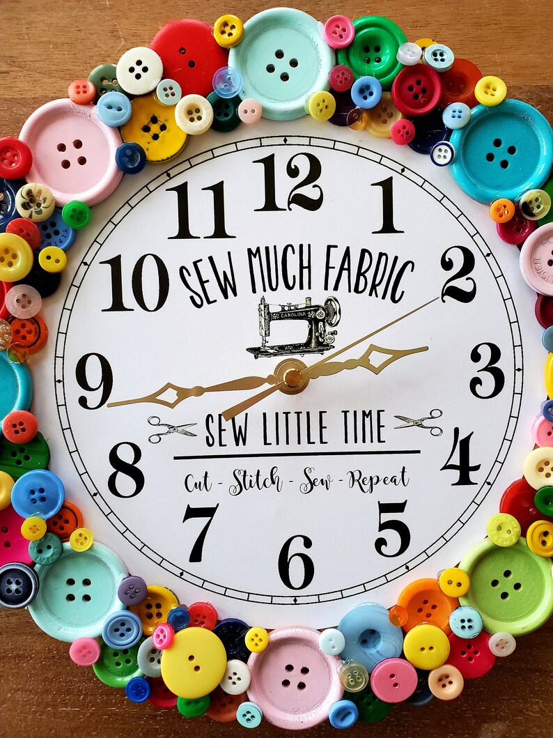May include: A white clock with black numbers and a gold hand. The clock is decorated with colorful buttons and the text "10 Sew Much Fabric 2 Sew Little Time Cut - Stitch - Sew - Repeat".