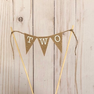 Puede incluir: Un adorno para tarta con forma de bander&iacute;n r&uacute;stico de arpillera con la palabra "TWO" en letras blancas. El bander&iacute;n est&aacute; ensartado en hilo y sujeto a dos palillos de madera, ideal para una celebraci&oacute;n de segundo cumplea&ntilde;os.
