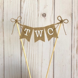Puede incluir: Adorno para tarta con una pancarta de arpillera con la palabra "TWO" en letras blancas. La pancarta est&aacute; suspendida entre dos palitos de madera, adornada con lazos de cuerda. Sobre un fondo de madera clara.