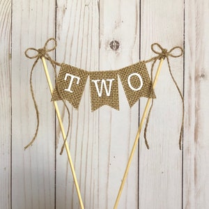 Puede incluir: Un adorno para tarta con forma de bander&iacute;n de arpillera con la palabra "TWO" en letras blancas. El bander&iacute;n est&aacute; ensartado en clavijas de madera y atado con lazos de cuerda. Perfecto para una celebraci&oacute;n de segundo cumplea&ntilde;os.