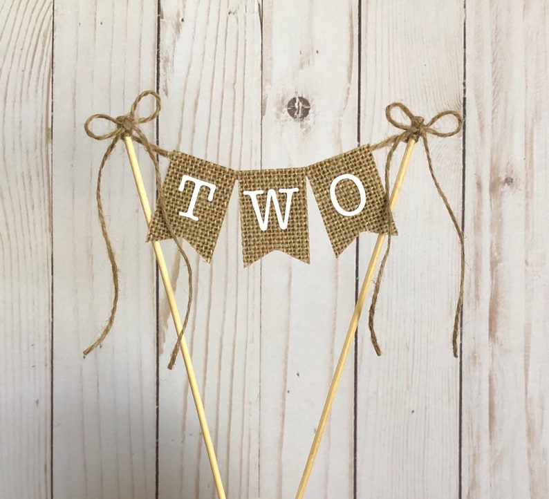 Puede incluir: Un adorno para tarta con forma de bander&iacute;n de arpillera con la palabra "TWO" en letras blancas. El bander&iacute;n est&aacute; ensartado en clavijas de madera y atado con lazos de cuerda. Perfecto para una celebraci&oacute;n de segundo cumplea&ntilde;os.