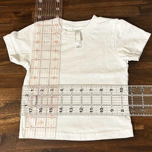 May include: A white t-shirt with a ruler measuring the length and width of the shirt. The ruler shows measurements in inches.