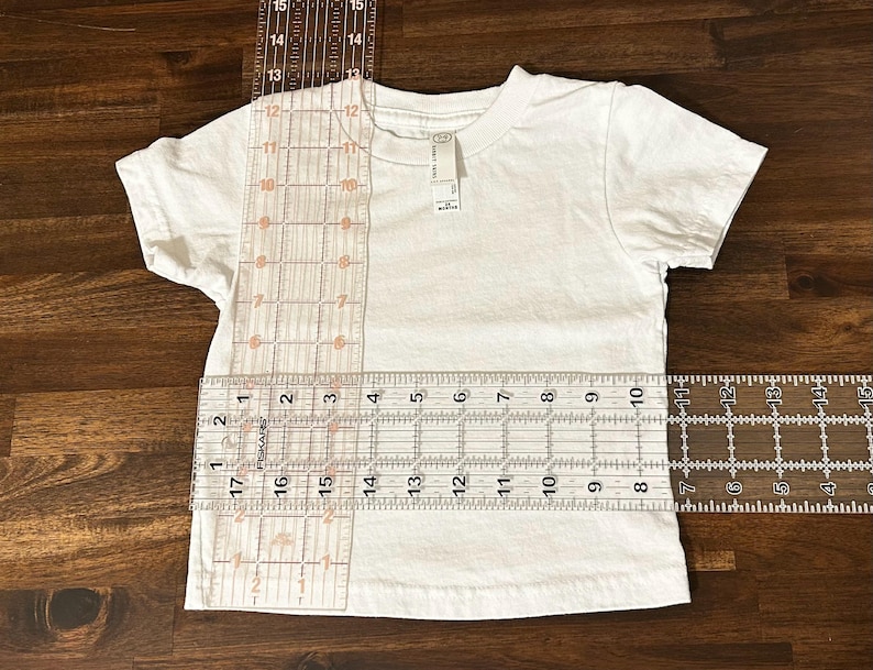 May include: A white t-shirt with a ruler measuring the length and width of the shirt. The ruler shows measurements in inches.