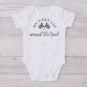 May include: White baby onesie with black text that reads "My First Lap Around The Track" with a checkered flag graphic.